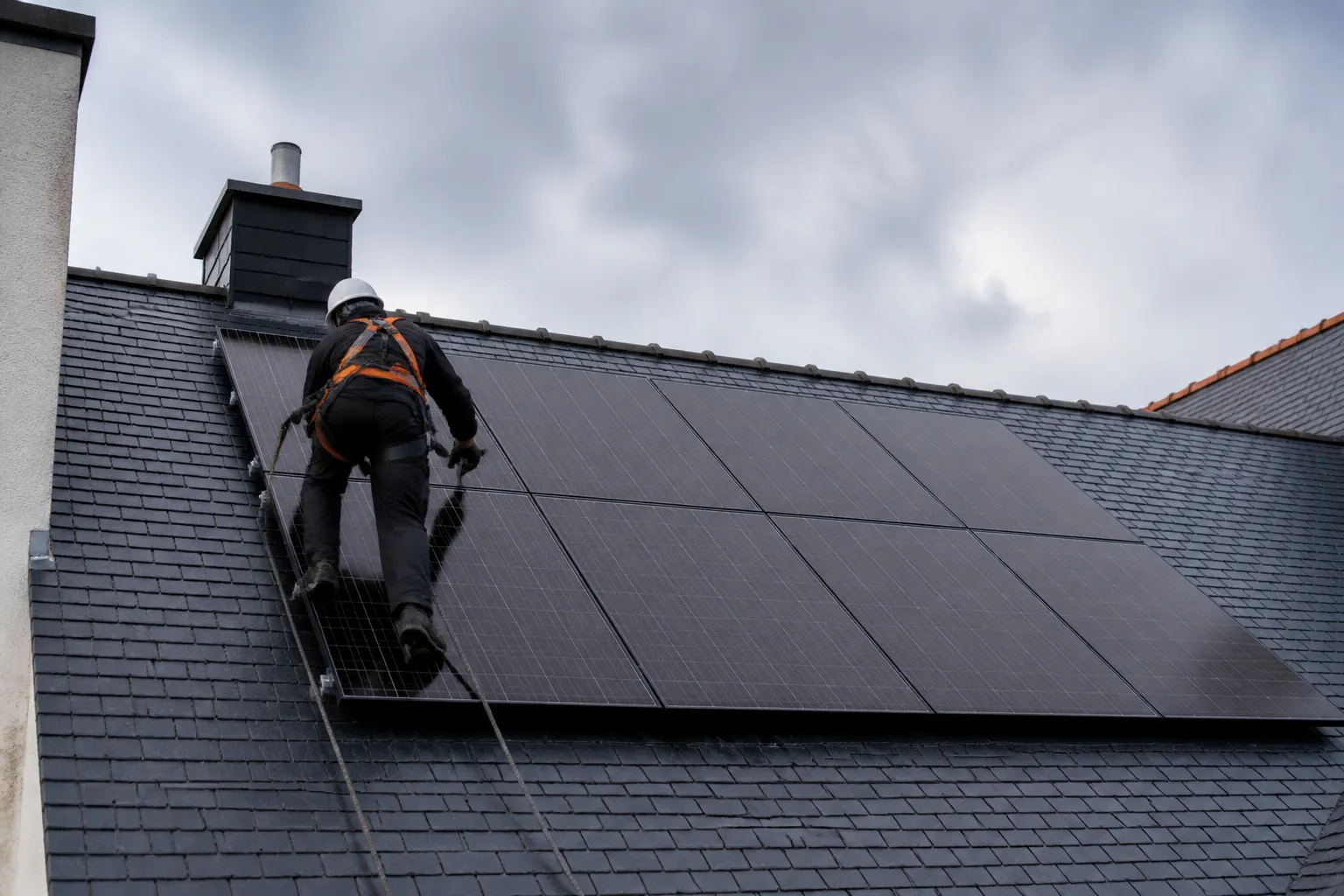 Installateur en harnais de sécurité contrôlant un panneau solaire sur une toiture en ardoise, vu de dos